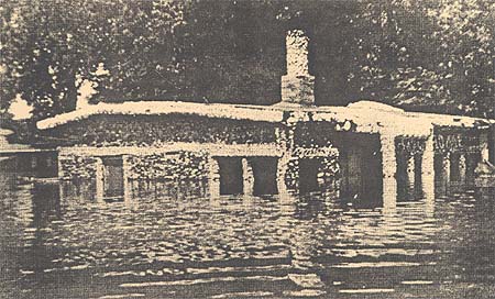 <b>Shell-covered House</b><br>This photograph, taken during the 1943 flood, shows a small, single-story house with one chimney, a gently pitched roof, numerous windows and an entryway covered by a small porch with two vertical support posts.  The outer surface of the structure is covered with dark and light-colored objects, presumably shells and perhaps some stones, which are clearly arranged in patterns.  Light-colored shells cover the upper portion of the chimney, all eaves, including those of the porch roof, the porch's support posts and some window frames.  A horizontal band of light shells extends around the structure just above the window openings.  Light-colored shells also spell out a sequence of letters above the row of windows appearing on the right-hand side of the photograph.  They appear to spell out 