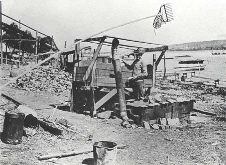 <b>Musseler's Camp</b><br>A square tub on blocks, used for boiling the mussels, can be seen in front with shells lying on it.  Two shell forks and clam dredge with a very long handle are leaning on a table used when cleaning the mussel meat out of  the shells.