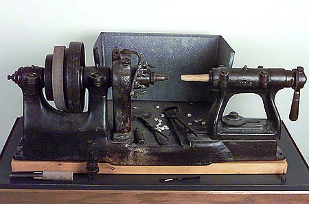 <b>Button Cutting Machine and Tools</b>.<br>The shell pusher is to the right, the saw surrounded by water jets is in left-center, and the pulley that holds the belt to turn the saw is on the left.  Lying in the bed of the cutter are a small hammer used to tap cut blanks out of the shell and shell tongs.<br>Jake Wolf Fish Hatchery exhibit, Topeka, Illinois.