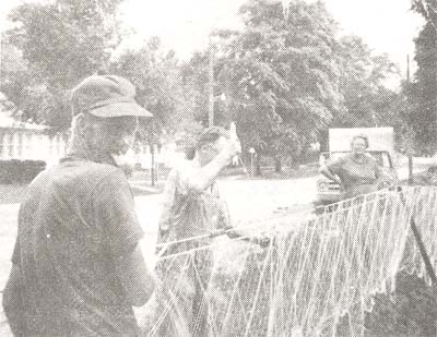 <b>Sam Hall Repairs a Trammel Net</b> using a net needle.  His wife and son are watching.